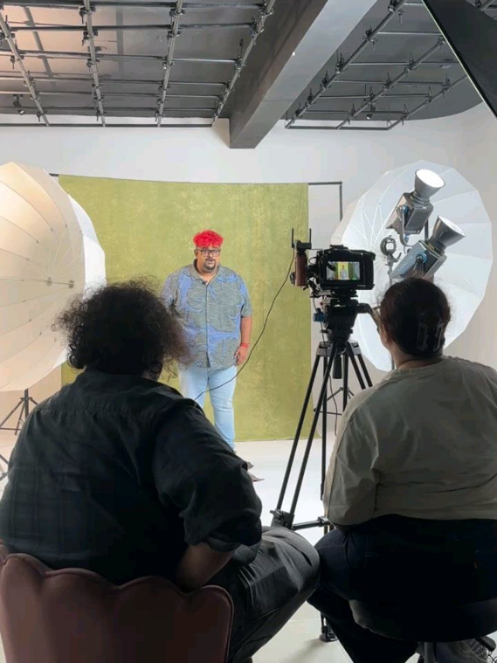 Man with bright red hair in a blue shirt stands before a green screen while being filmed in a studio set with lighting and a camera on a tripod.
