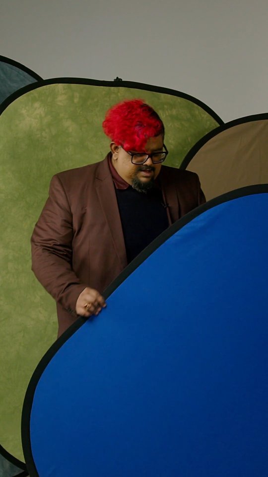 Man with bright red hair and glasses in a brown blazer, standing beside large blue and green photography backdrops in a studio environment.