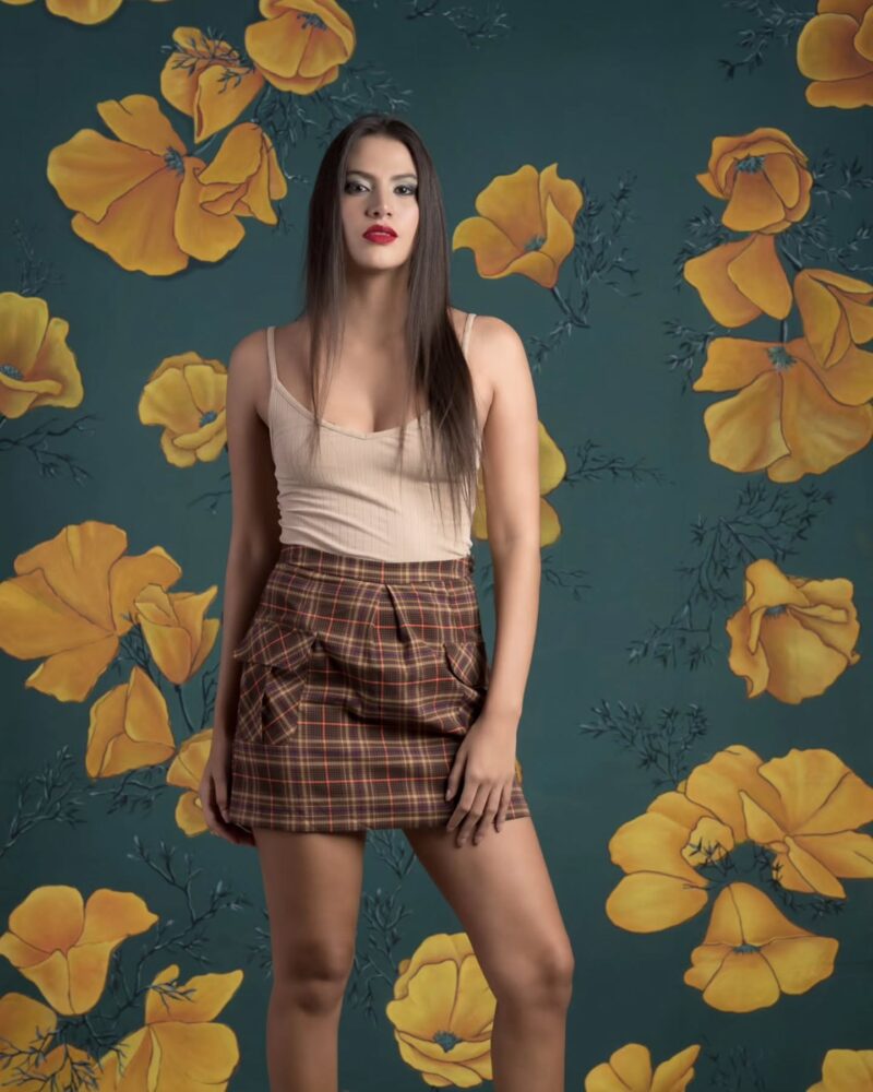 Woman posing in a beige tank top and brown plaid skirt, standing before teal wallpaper with large yellow flowers.