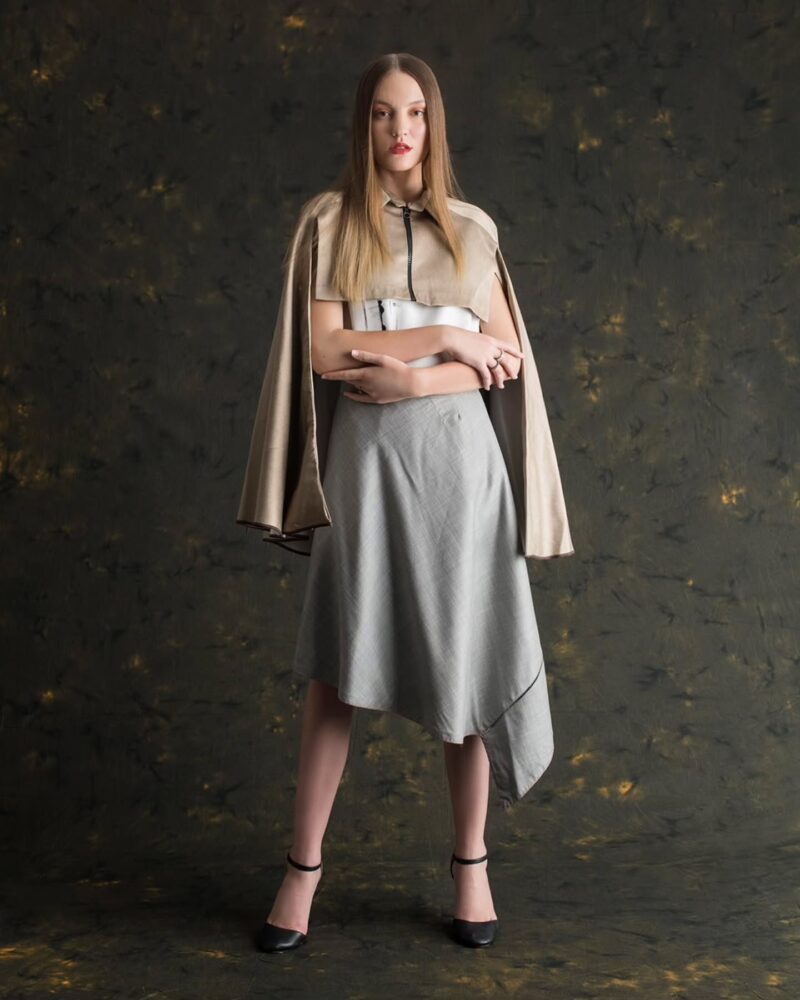 Full-length fashion shot of a woman wearing a beige cape-style outerwear over a light blouse and gray A-line skirt, standing with arms crossed in front of a dark textured backdrop.