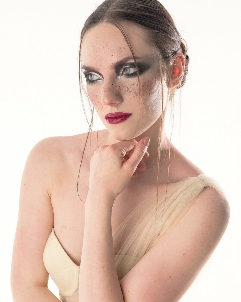 Portrait of a woman with bold smokey eye makeup, red lips, and freckles, wearing a beige strapless top and looking to the side.