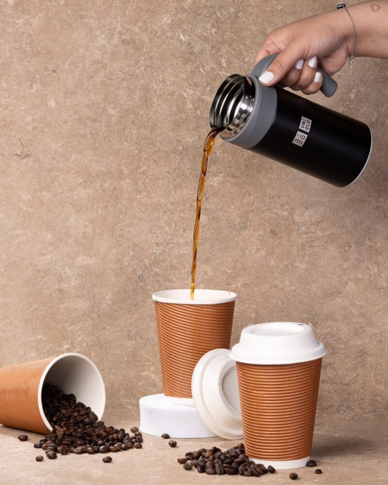 Hand pours hot coffee from a black insulated tumbler into a brown paper cup, with scattered coffee beans on a textured surface near the bottom left.