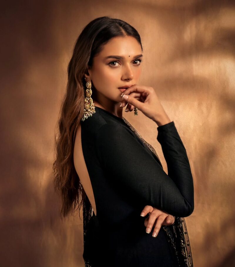 Portrait of a woman in a backless black gown with elaborate gold earrings, posing against a warm bronze backdrop.