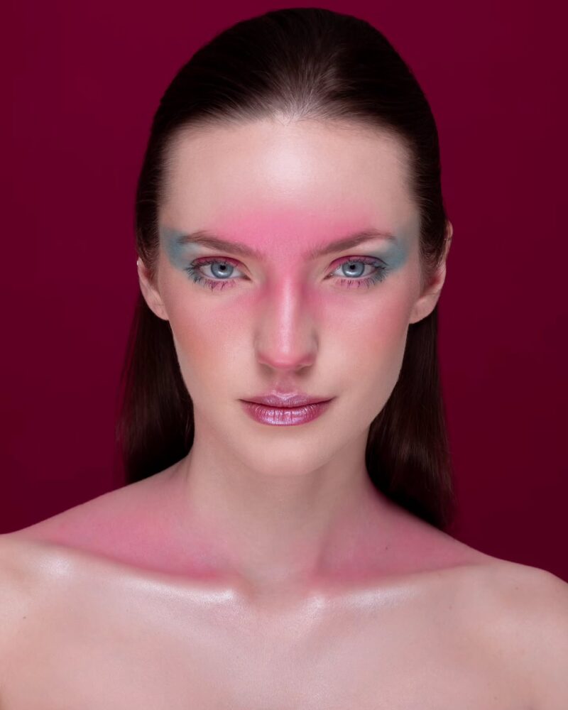 Close-up portrait of a woman with bold pink eye makeup and blue eyeshadow against a burgundy background.