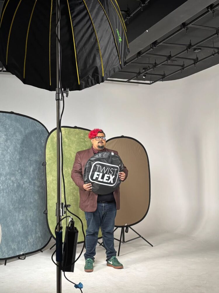 Man in a brown blazer holding a large Twist Flex collapsible backdrop, in a photography studio with lighting and color backdrops behind him