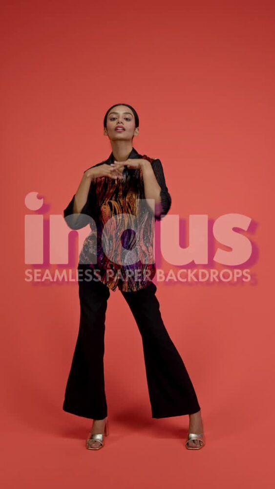 Female model standing in a coral studio backdrop, wearing a tiger-print blouse, black wide-leg pants, and silver heels, posing with hands near her chest; a pink watermark overlays the image.