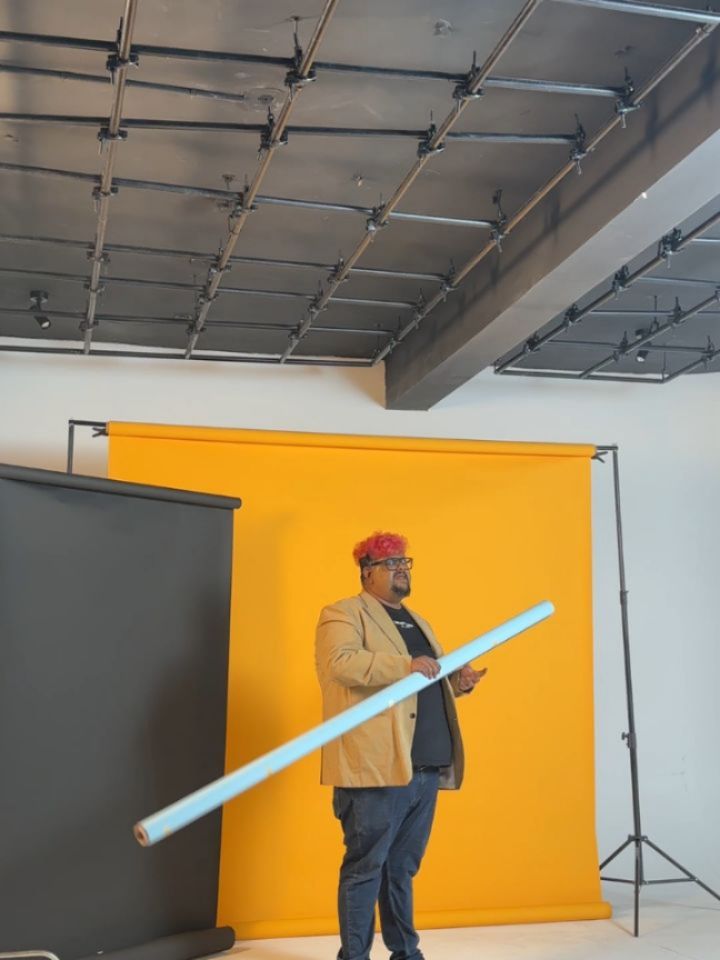 Man with pink hair and glasses posing in a studio in front of a yellow backdrop, holding a light blue rolled poster tube.