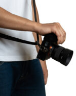 Person's hand grips a black camera with a strap, wearing a white shirt and blue jeans against a white background