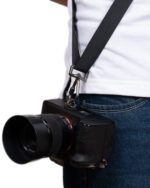 DSLR camera with a large lens and black strap hanging from a person wearing a white shirt and blue jeans, close-up at the waist area.