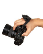 Close-up of a hand gripping a black professional camera with a wide lens and a strap looped around the wrist, photographed from the side against a white background.