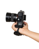 Hand holding a black DSLR camera with a large zoom lens, against a white background, with a camera strap beneath the hand.