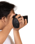 Person with short dark hair holding a black camera up to their eye, ready to take a photo against a white background.